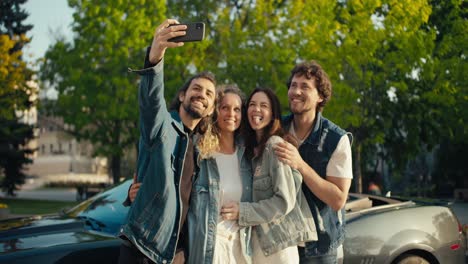 un grupo feliz de amigos vestidos con chaquetas de mezclilla toman una selfie frente a un descapotable gris en un soleado día de verano