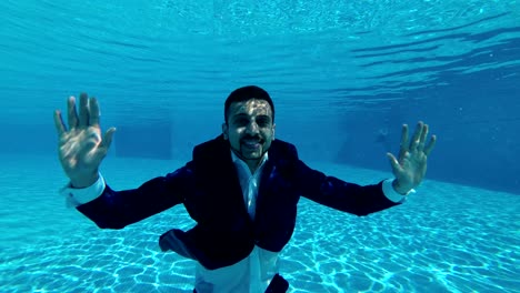 a man in a blue suit and a white shirt swims and poses underwater in the pool. he looks at the camera, waves his hands and smiles.