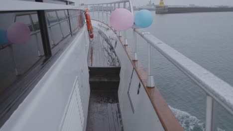 walking along starboard side of yacht on rainy day, traveling towards harbor, pink and blue balloon, luxury gender reveal party