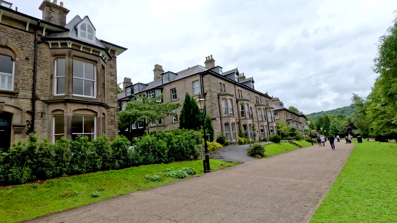Camera moves smoothly along a tree-lined path beside Victorian stone buildings in Buxton, England, under overcast daylight, capturing greenery and pedestrians