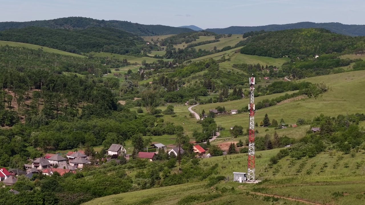 Un traliccio per telefonia mobile si erge imponente in un vasto paesaggio verde, dominando un piccolo villaggio rurale sotto un cielo estivo sereno