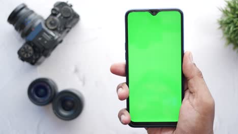 Top-view-of-holding-smart-phone-with-green-screen-and-camera-on-background-,