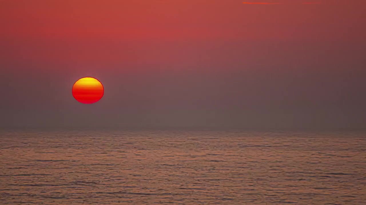Setting red and yellow sun dips below horizon of ocean on clear sky day
