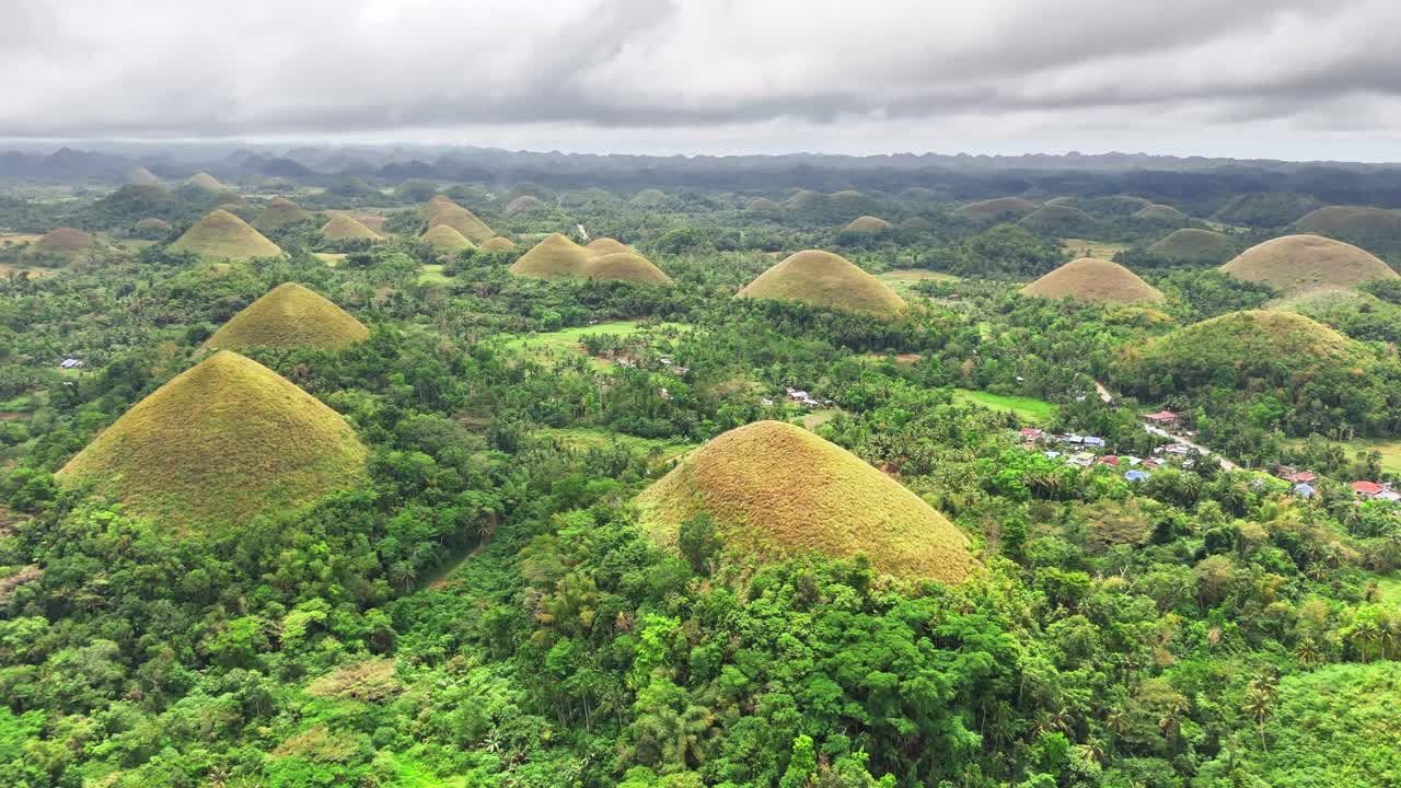Aerial landscape of the Chocolate Hills in Bohol, Philippines, where hundreds of cone-shaped grass hills rise from a dense tropical forest, creating a stunning natural terrain beneath a cloudy sky