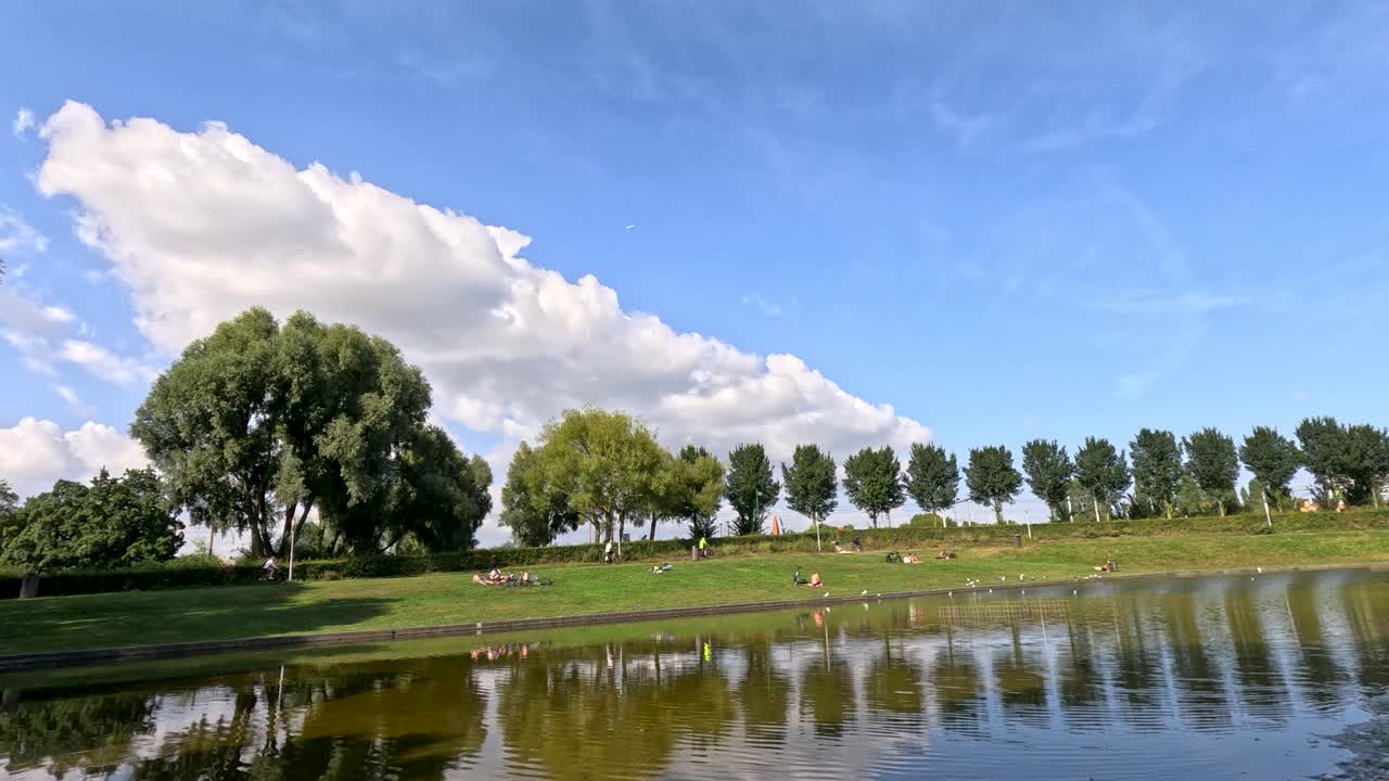 Wide daylight pan across Haarlem park, green grass, trees, blue sky, pond reflections, relaxed atmosphere