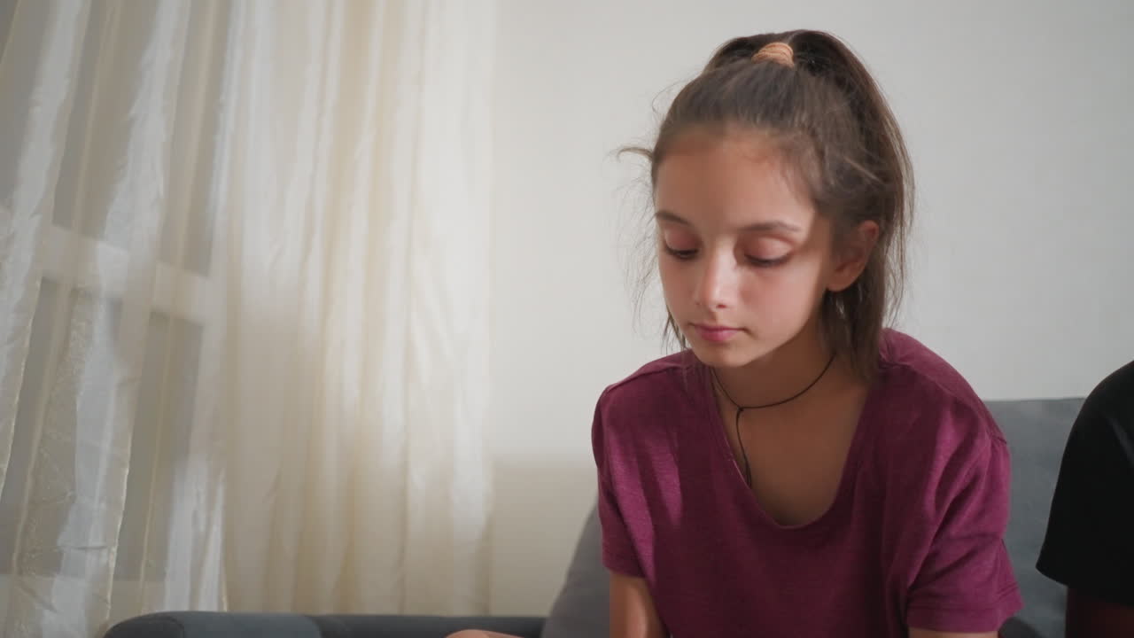 Chica con camisa burdeos sentada en el interior mirando hacia abajo con expresión concentrada mientras una luz suave se filtra a través de una cortina transparente en un ambiente hogareño tranquilo con una vista parcial de alguien sentado a su lado en un cómodo sofá.