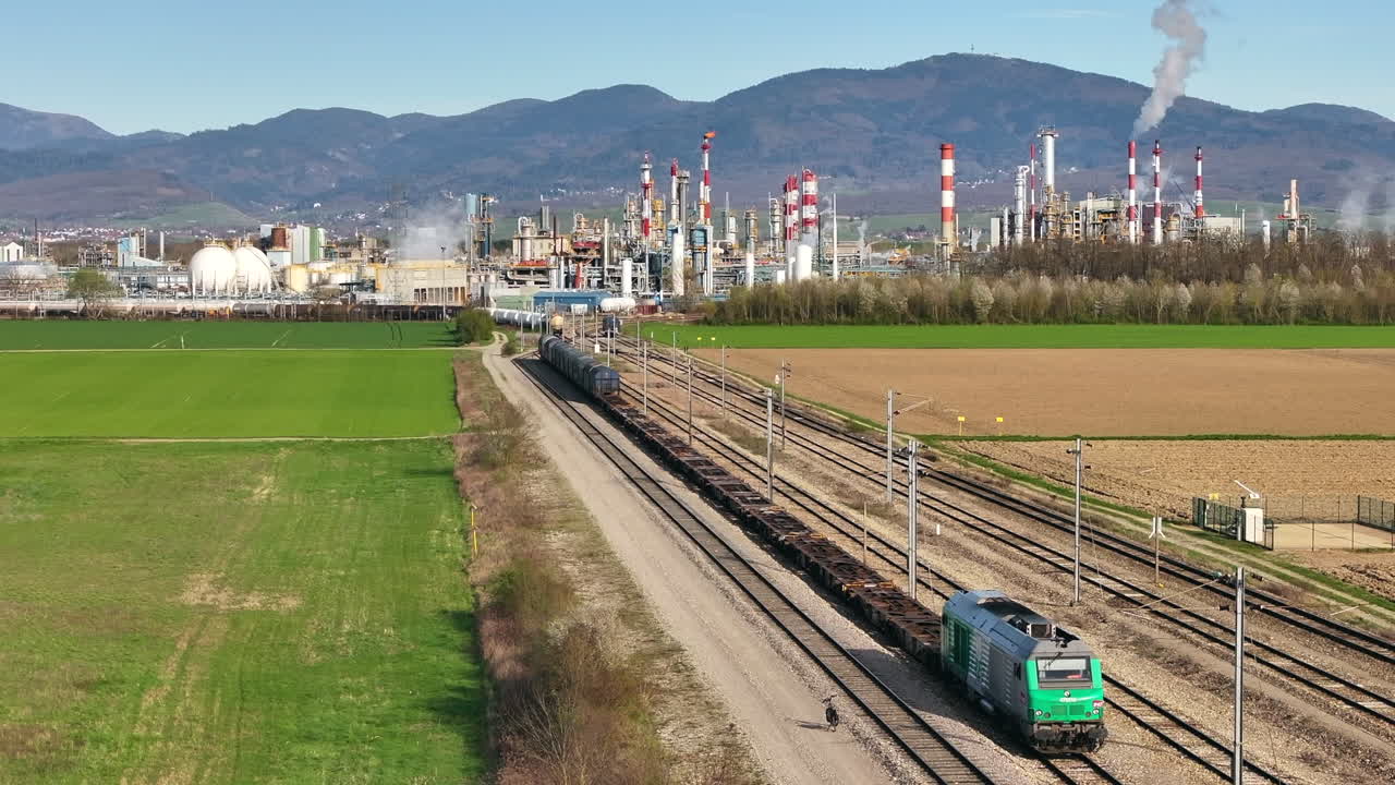Green locomotive on railway with Alsachemie factory in background, sunny day, industrial scene