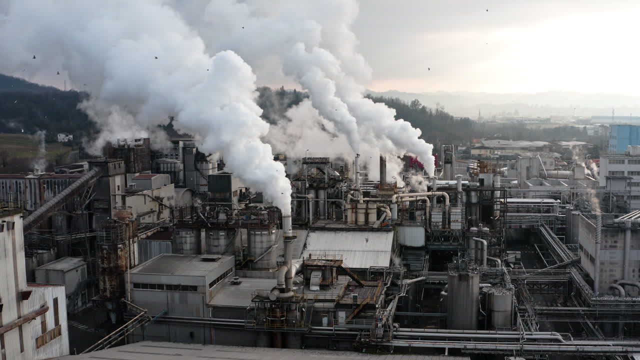 Industrial factory emitting thick white smoke from multiple chimneys under a cloudy sky