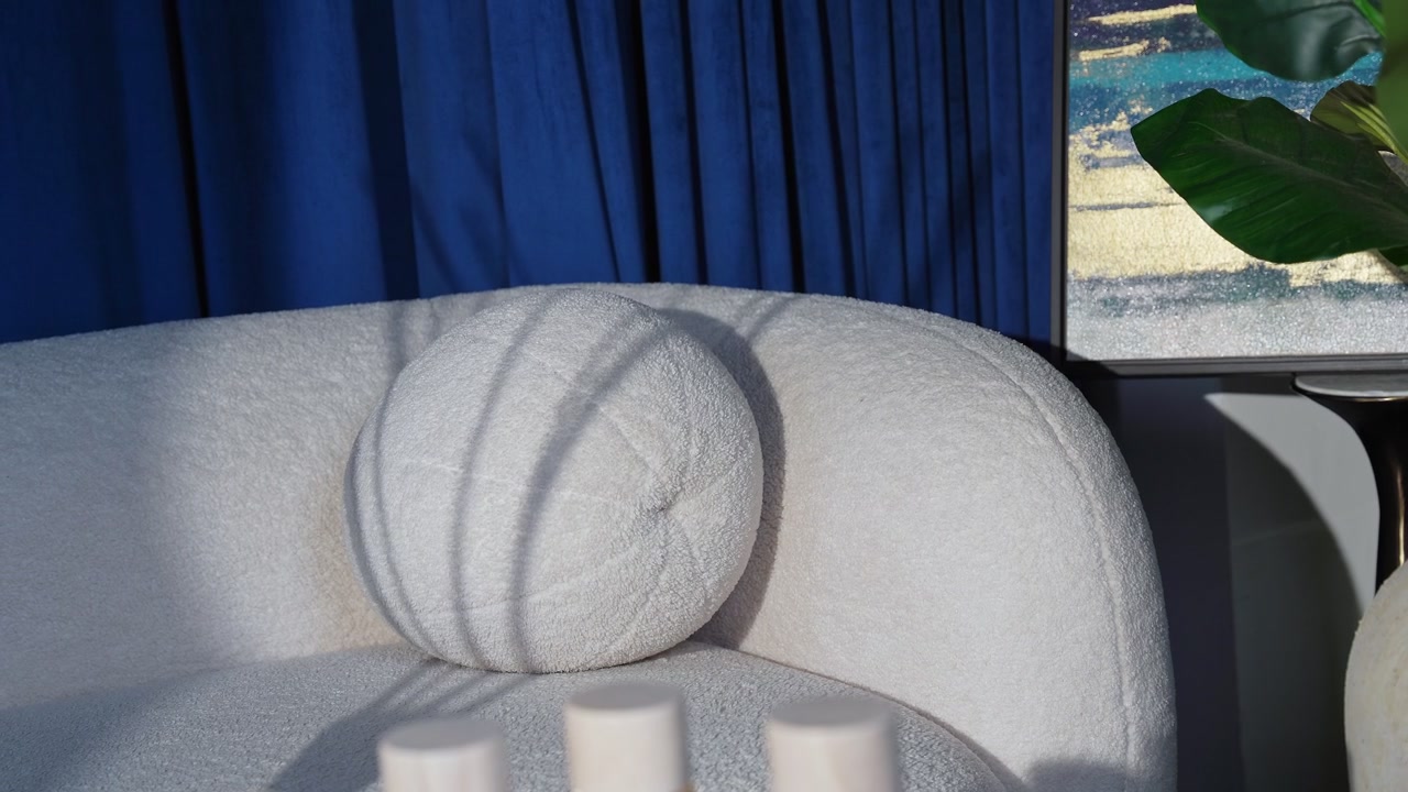 Un'inquadratura cinematografica di un lussuoso divano bouclé bianco e un cuscino a sfera con ombre drammatiche dalle finestre, con tende di velluto blu intenso e arte astratta in un interno moderno.