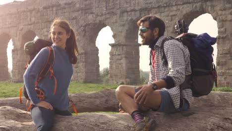 happy young couple backpackers tourists sitting on a log trunk talking and smiling in front of ancient roman aqueduct ruins in parco degli acquedotti park in rome at sunrise with guitar sleeping bag slow motion steadycam