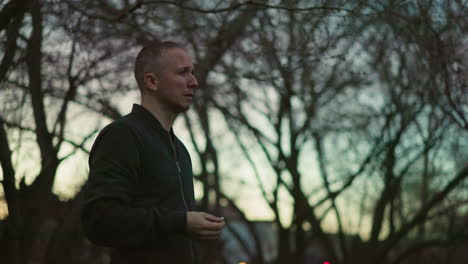 a side view of a man in a green jacket smoking a cigarette in the forest at dusk, with blurred background of trees and the dimming light