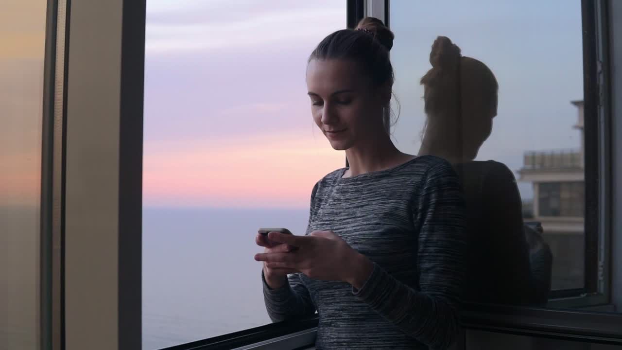 hermosa joven sentada en casa junto a la ventana abierta con una sonrisa mirando el teléfono y escribiendo un mensaje durante el atardecer