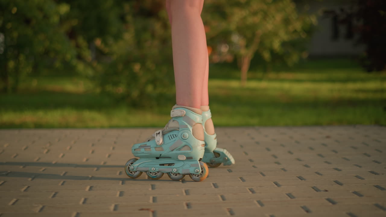 Side leg view of person roller skating on interlocked path, wearing teal rollerblades, shadow cast on ground, next to grassy field, blurred background of greenery