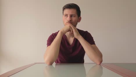 Young-man-sitting-at-table-being-indecisive-about-a-choice-and-thinking-hard,-medium-close-up-shot-facing-camera-indoors-with-natural-light