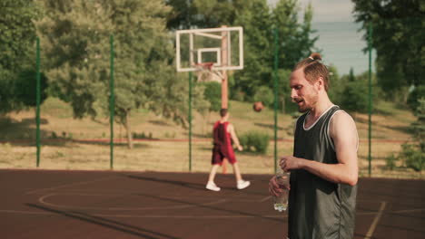 In-Foreground,-A-Handsome-Blonde-Bearded-Basketball-Player-Taking-A-Break-And-Drinking-Water-In-An-Outdoor-Basketball-Court