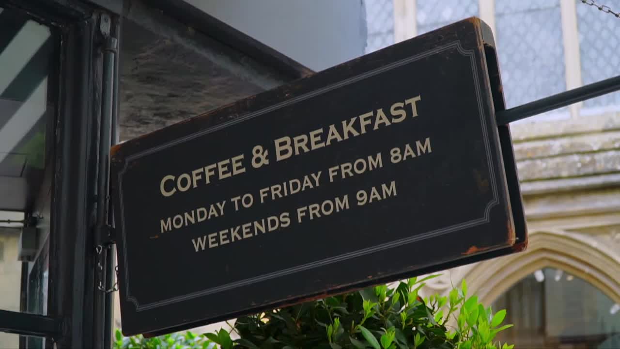 Close-up of an old café sign displaying coffee and breakfast hours, hanging outside a shop in the historic center of Salisbury, England.
