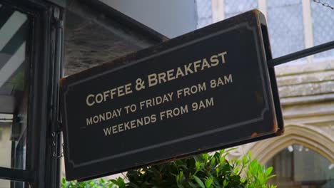 close-up of an old café sign displaying coffee and breakfast hours, hanging outside a shop in the historic center of salisbury, england.