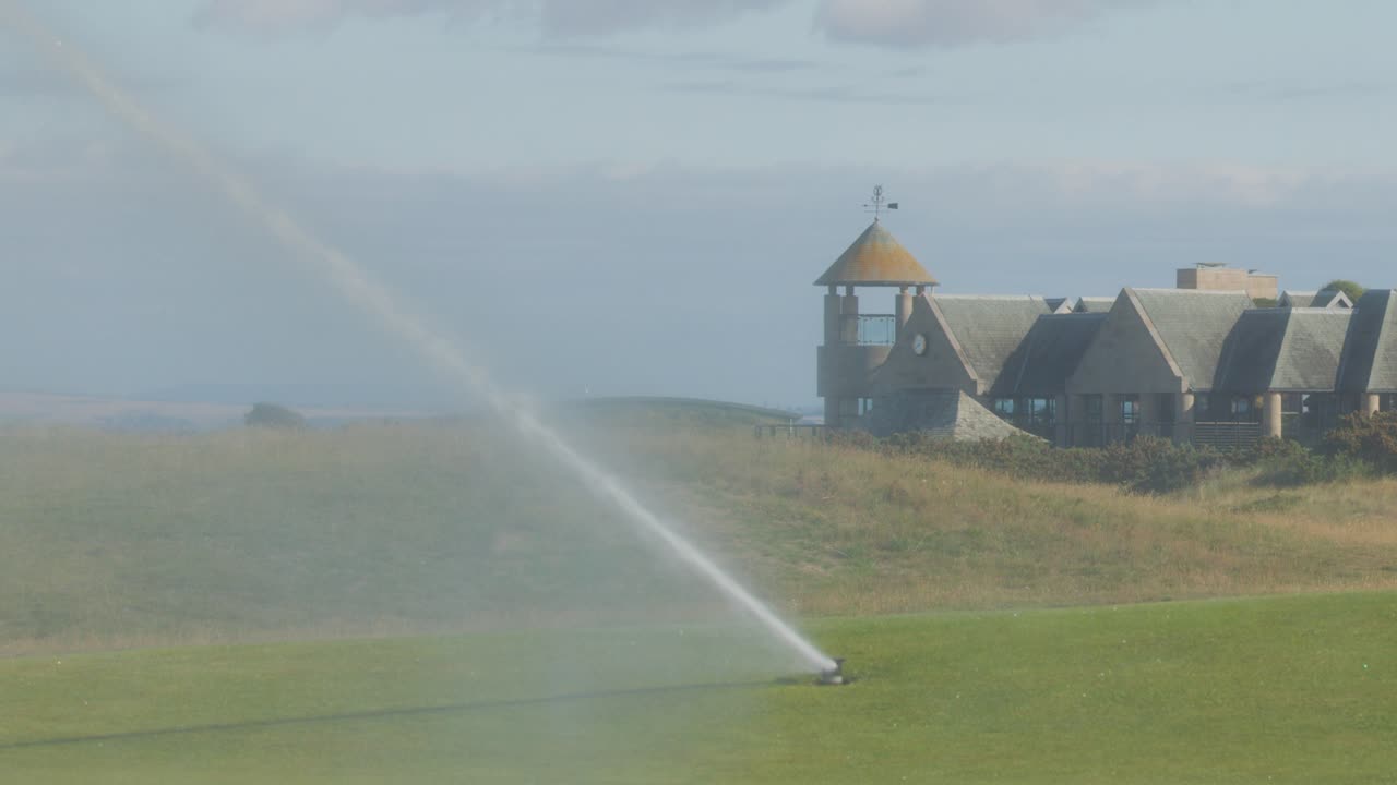 A rotating sprinkler irrigates a green golf course under daylight, with a clubhouse and distant hills in the background. Wide, steady shot with natural lighting