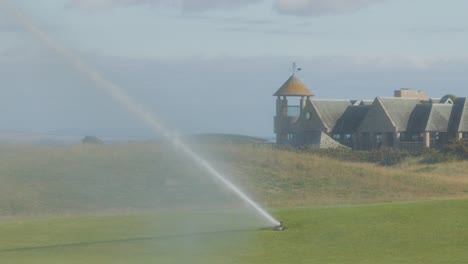 a rotating sprinkler irrigates a green golf course under daylight, with a clubhouse and distant hills in the background. wide, steady shot with natural lighting