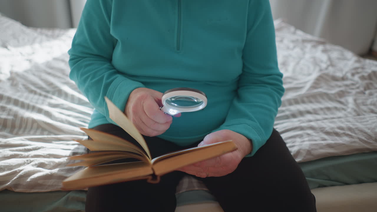 Older woman using magnifying glass to read book. Sitting comfortably in cozy bedroom, focused on text with light shining on the page, highlighting details while enjoying peaceful reading moment