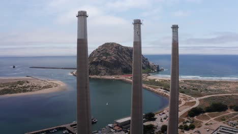 Close-up-aerial-dolly-shot-of-the-famous-three-smokestacks-of-the-Morro-Bay-Power-Plant-with-Morro-Rock-in-the-background-in-Morro-Bay,-California