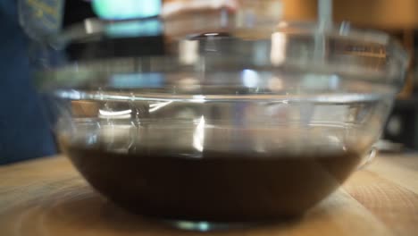 chef is adding a cup of soy sauce into a clear glass bowl along side a variety of other liquid and dry ingredients.