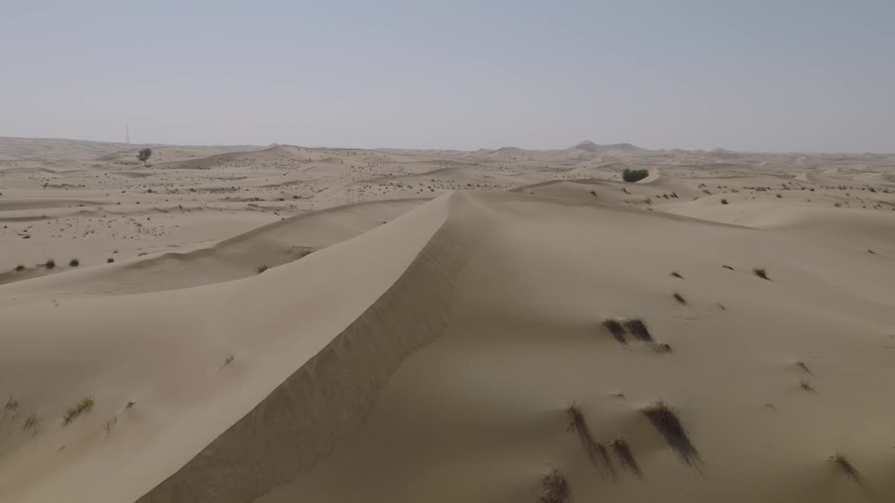 Ripresa cinematografica con drone che si muove a destra, ascendendo e ruotando sottilmente per mantenere come fuoco la cresta affilata e curva di una duna di sabbia. Il movimento rivela l'ampio e aperto deserto.