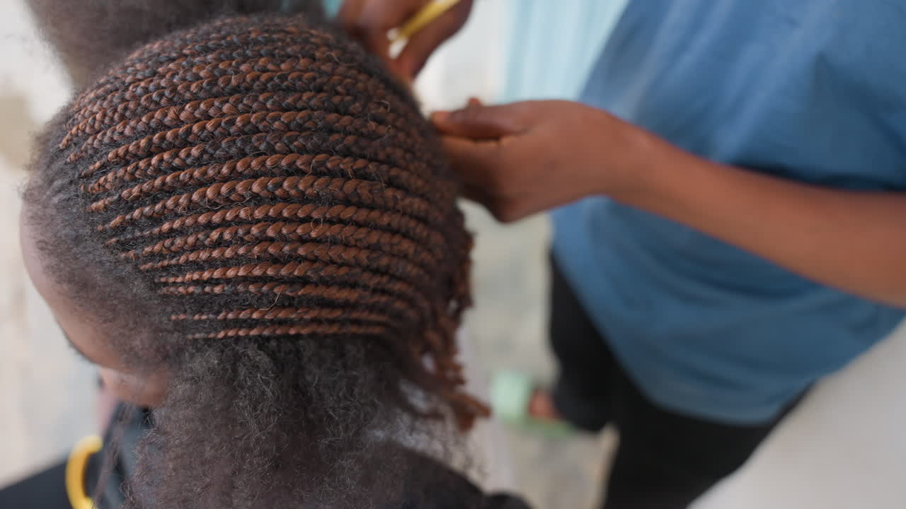 Styliste noire tressant des tresses africaines soignées, gros plan en plongée sur des mains expertes formant des rangées précises sur des cheveux texturés, atmosphère de studio concentrée, lumière naturelle mettant en valeur les tons bruns et les détails du cuir chevelu
