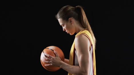 Una-Joven-Caucásica-Enfocada-Sostiene-Una-Pelota-De-Baloncesto-En-Un-Ambiente-Oscuro-Sobre-Un-Fondo-Negro,-Transmitiendo-