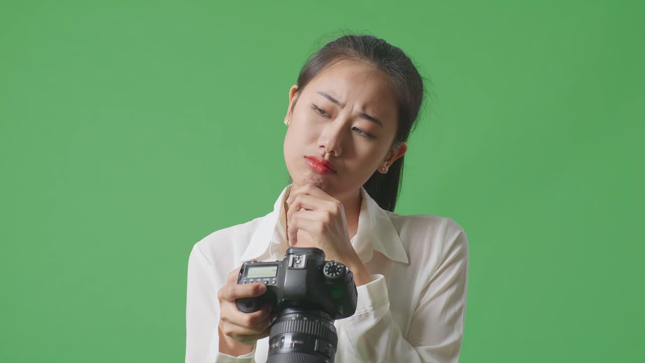 아시아인 사진작가가 사진과 영상에 대해 생각하고, 스튜디오에서 녹색 스크린 배경에서 카메라에서 사진을 보는 동안 지표손가락을 들어 올립니다.