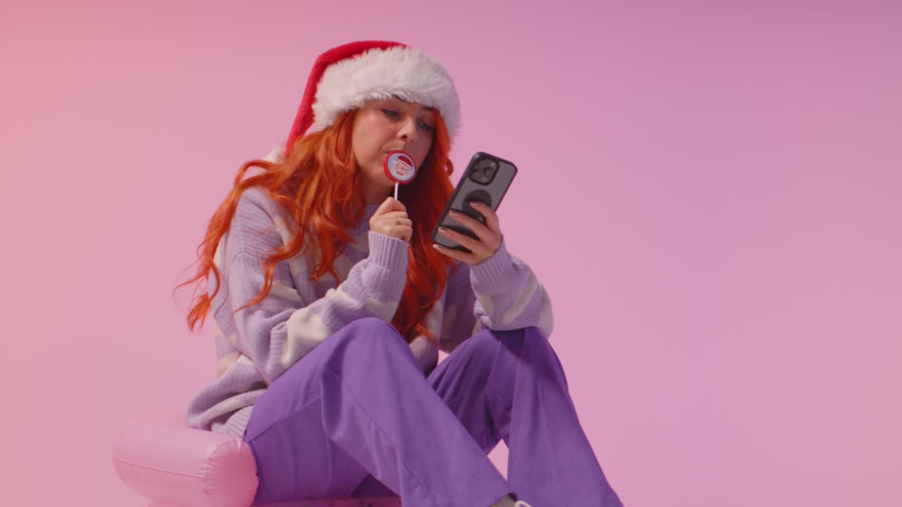 fotografía de estudio de una mujer joven de la generación z con sombrero de papá noel comiendo caramelos y mirando el teléfono móvil 2