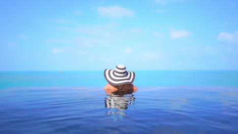 a woman in a floppy sun hat with her back to the camera looks out from her resort swimming pool at the ocean beyond