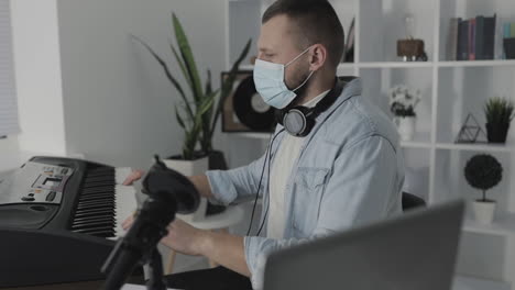 músico masculino con mascarilla médica tocando teclado eléctrico en casa durante el encierro debido a la pandemia de covid 19