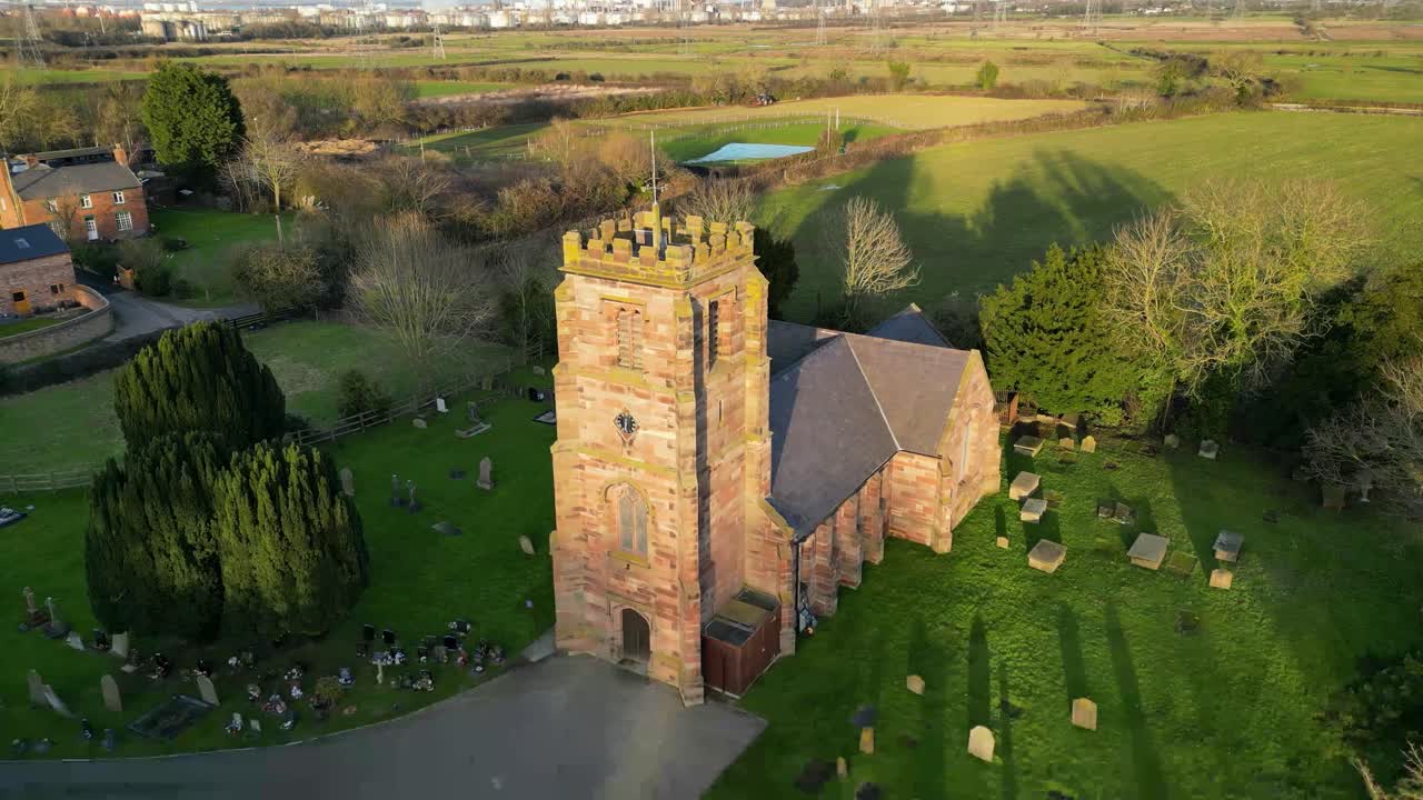Aerial view of historic stone church with clock tower and surrounding graveyard in rural Hampshire, England, showing warm sunlight over green countryside and traditional village setting