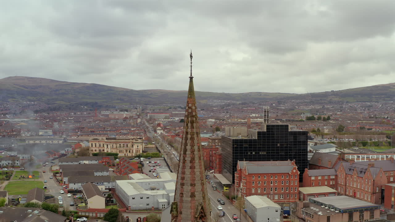 Dramatic aerial orbit of Gothic church spire punctuating Belfast's moody cityscape beneath overcast skies