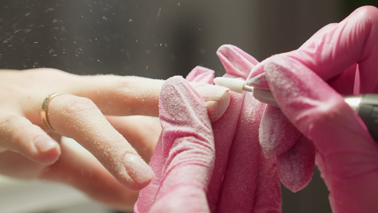 Close-up view of nail technician in pink gloves shaping client nail using electric file. Fine dust scatters in air as client s hand with gold ring is gently held for detailed manicure work