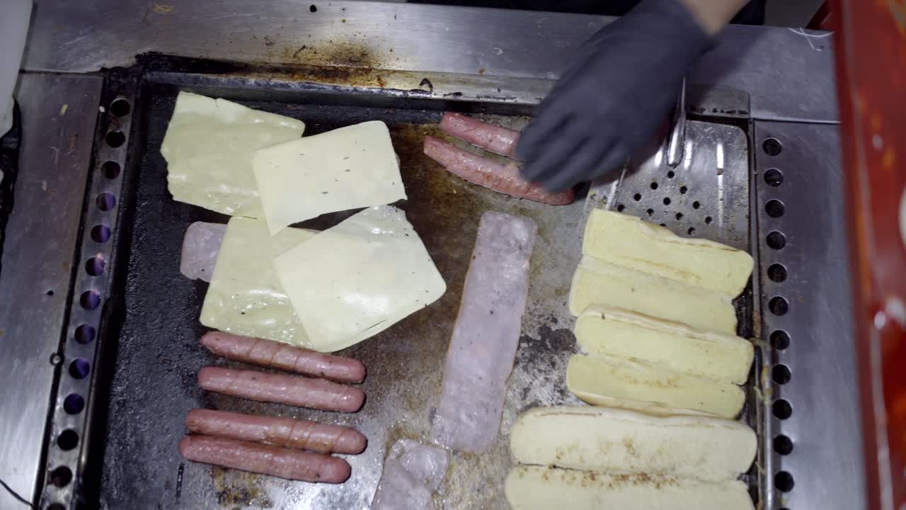 Zenithal shot of cheese slices being placed on sausages cooking on a flat top grill in a street food stand