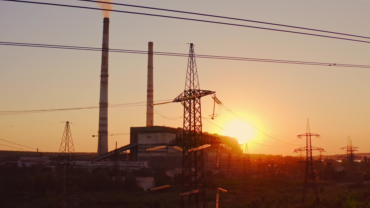 Electricity pylons at sunset. High-voltage electric tower on industrial pipe background. Transmission lines near the factory with dirty smoke into the air in the evening.