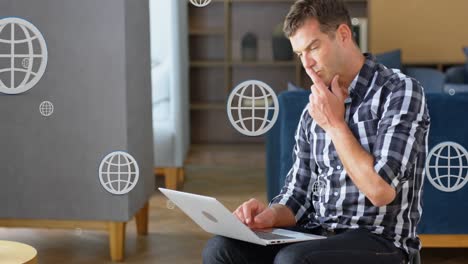 Animation-of-multiple-globe-icons-over-caucasian-man-sitting-on-couch-and-working-on-laptop