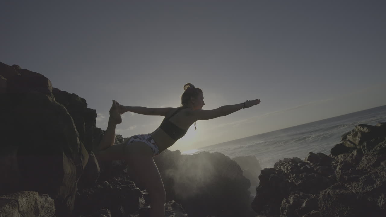 Woman practicing yoga on a rocky coastline at sunrise
