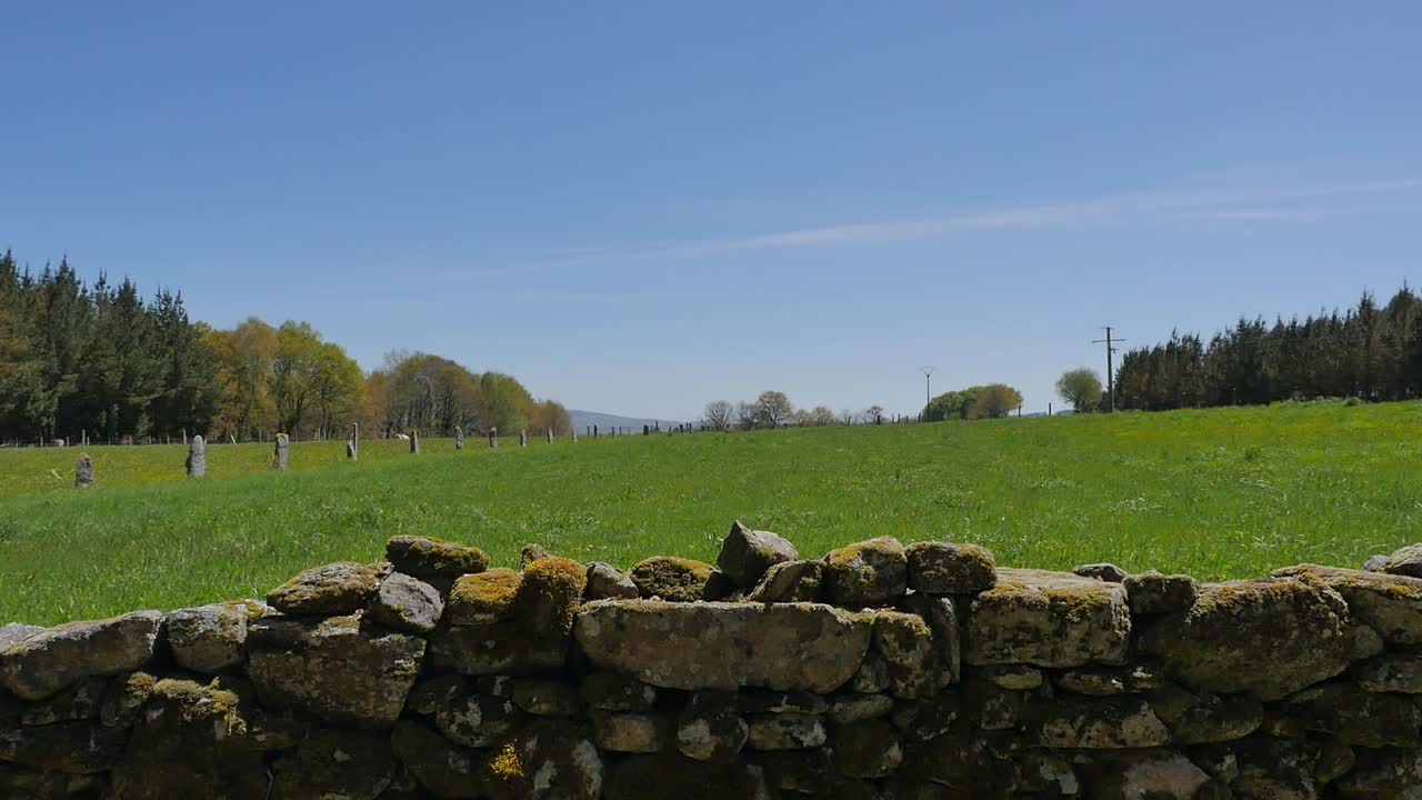 un campo de hierba abierto con un muro de piedra en primer plano y un bosque a cada lado, con un fondo de cielo azul en un día soleado