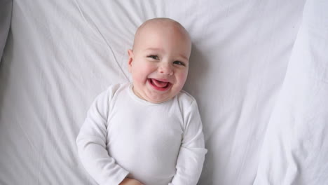 A-child-in-a-white-t-shirt-lying-on-a-white-bed-looking-at-the-camera-and-laughing-in-slow-motion,-top-view