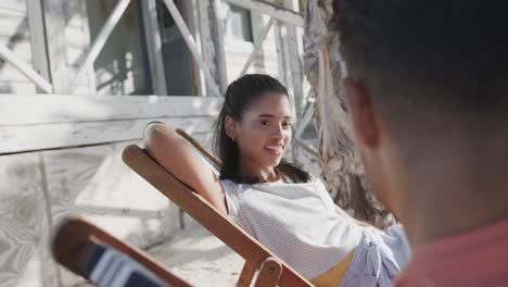 Happy-diverse-couple-sitting-in-deckchairs-talking-outside-wooden-beach-house,-in-slow-motion