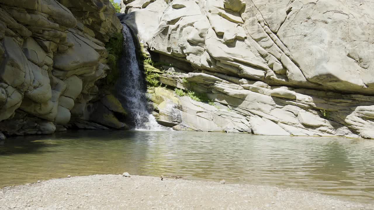 A steady waterfall cascades down smooth canyon walls into a calm, clear pool surrounded by sculpted rock formations.