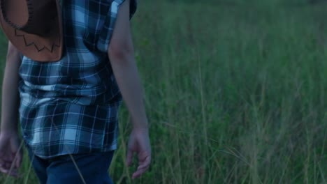 slow-motion follow shot of a cowgirl wearing a flannel shirt and cowboy hat walking through a grass field in moody setting