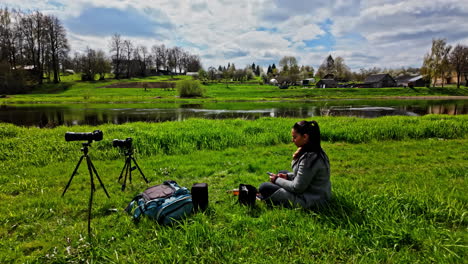 Woman-in-grey-long-coat-sits-along-river-banks-of-Bausta-watching-over-cameras-on-tripods