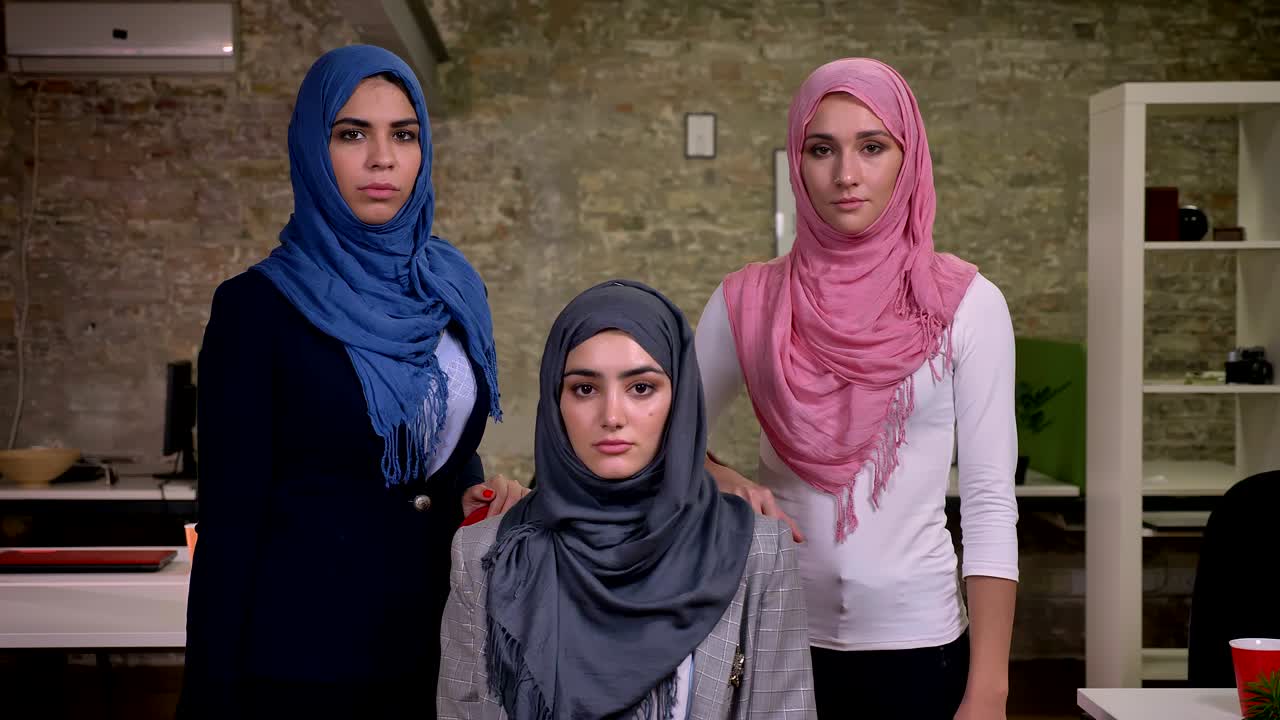 Tre allvarliga arabiska kvinnor står och tittar rakt in i kameran, bärande hijab, med en arbetsatmosfär från Mellanöstern, inomhus i ett modernt kontor med tegelväggar.
