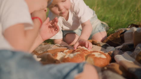 a close-up of two young boys wearing white tops, one lying down and the other bent over on a checkered picnic blanket in a grassy field. they are eating bread together