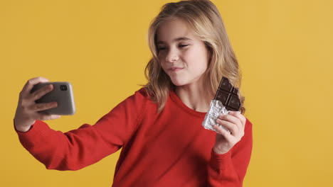 Teenage-Caucasian-girl-eating-chocolate-bar-and-taking-selfies-with-smartphone.