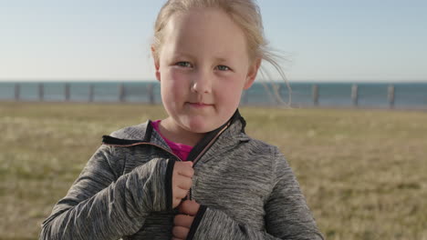 close up portrait of little blonde girl smiling at camera comfortable wearing cozy hoodie enjoying sunny seaside park